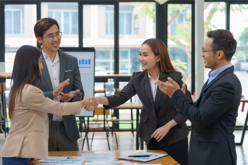 Four business professionals in formal attire are in a bright office. Two women in the center are shaking hands, while two men beside them are smiling and clapping, suggesting a successful meeting or agreement.