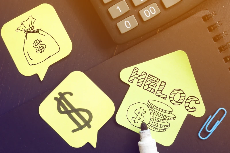 Yellow sticky notes with hand-drawn money symbols and “HELOC” inside a house shape, a calculator, a notebook, a marker, and a blue paperclip on a dark table.