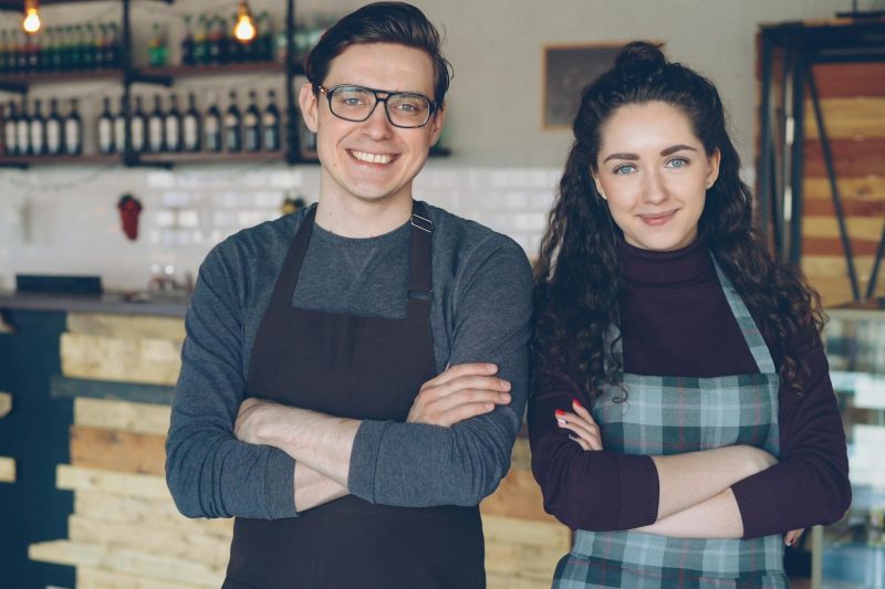 Two young cafe owners smile proudly while wearing aprons in their modern cafe setting.