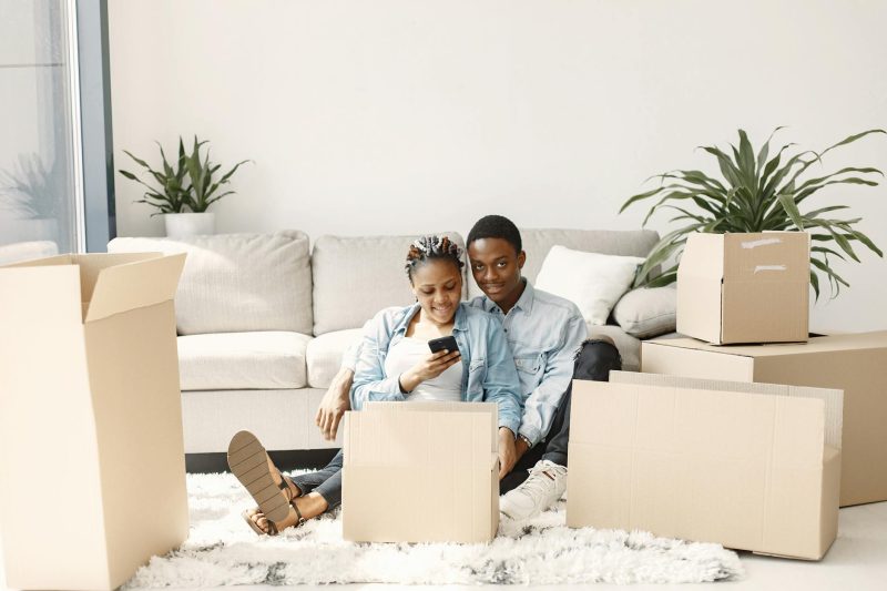 Happy couple sitting among cardboard boxes, smiling and using a smartphone to celebrate their new home.