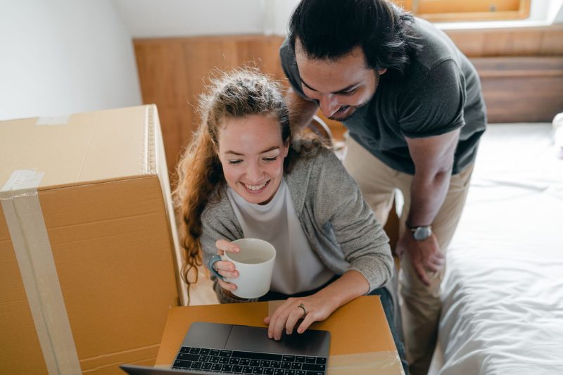 Cheerful woman typing on a netbook with a hot drink, while a man in a smartwatch leans forward in a new apartment surrounded by cardboard boxes.