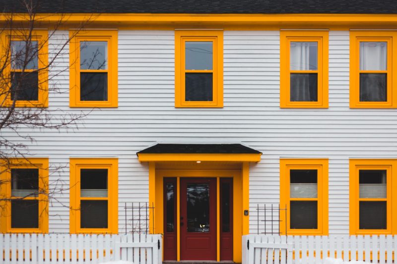 Vibrant yellow house with a red door, white fence, and multiple windows against a clear sky.